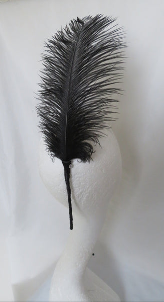A single black feather with a long stem, displayed on a white mannequin head against a white fabric backdrop.