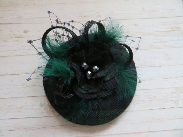 small round cocktail hat made from black green and blue blackwatch tartan with some feathers and a tartan flower black loops can be worn with clips or a headband matches the tartan dresses from british retro