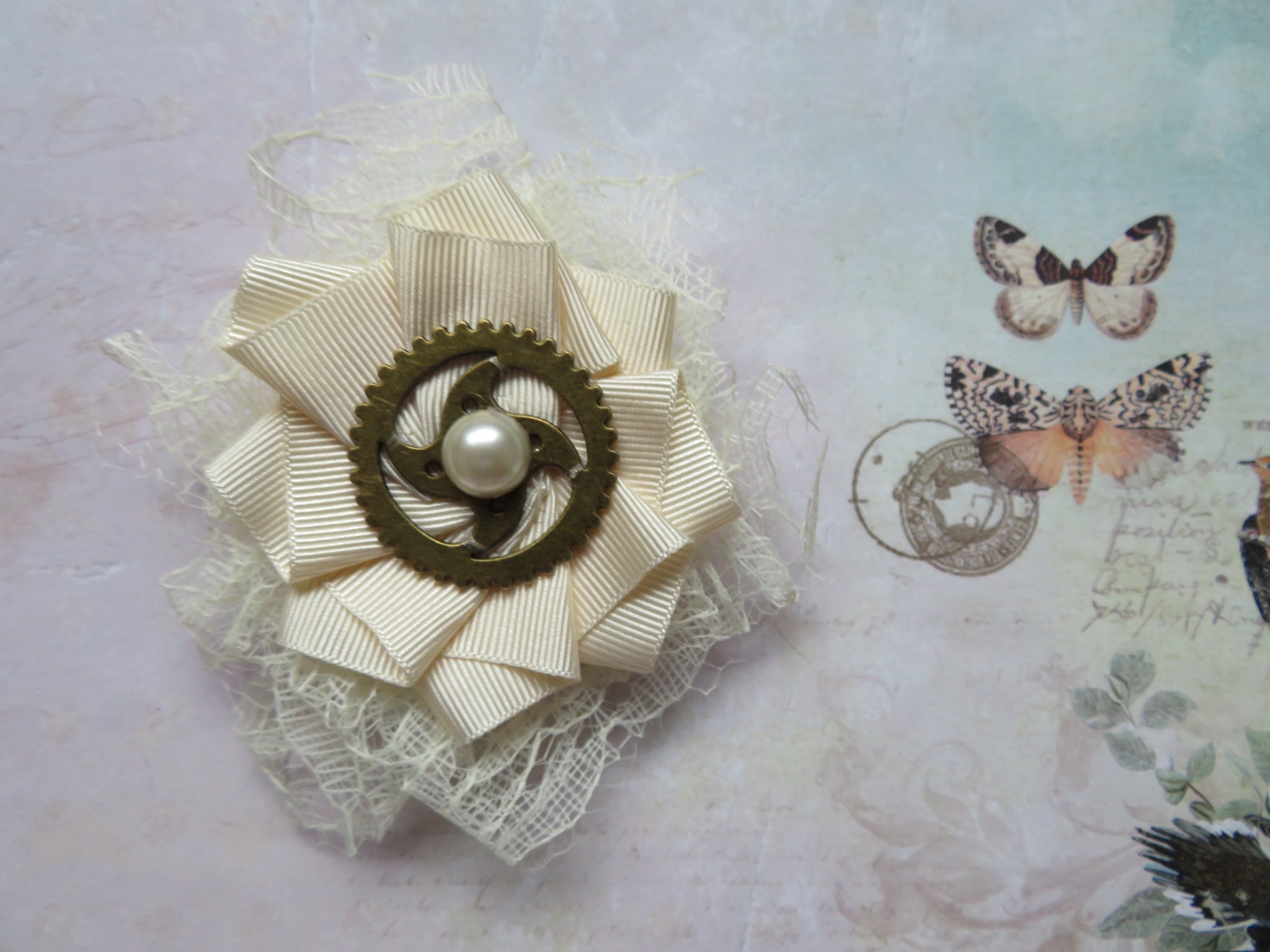 small brooch made with cream ribbed grosgrain ribbon handmade in a ruffle shape, with cream lace ruffle backing it large brass steampunk watch cog in the centre with a cream pearl in the centre to finish fixed with a roll pin brooch fixing at the back perfect for a steampunk wedding