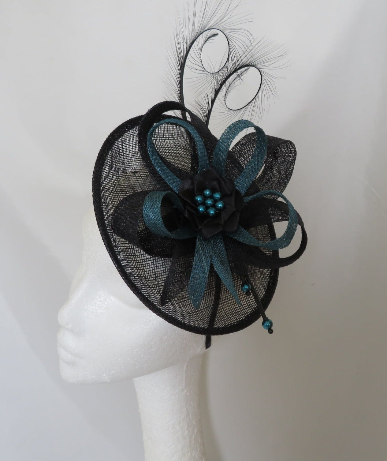 black and dark teal fascinator hat on a saucer base with an up lip black curled pheasant feathers and sinamay loops in wide and narrower in teal with a satin ribbon ruffle and teal pearls 