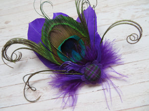 small feather hair or hat clip in dark purple with a peacock feather and fluffy feathers and a button at the bottom covered with purple pride of scotland tartan ribbon, on a hat clip