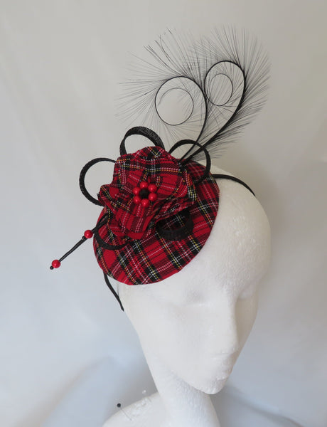 A red and black tartan plaid fascinator hat with black feather plumes and black ribbon loops, displayed on a white mannequin head against a white fabric backdrop.