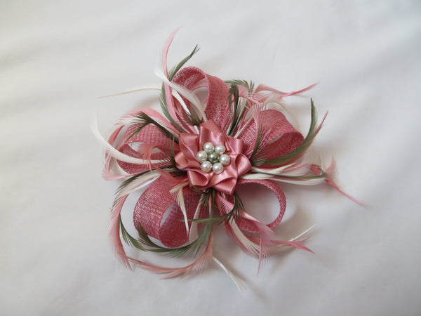 A pink fascinator hair accessory featuring a textured pink ribbon loop, a satin pink flower, and white feathers with a pearl-like center, displayed on a white fabric background.