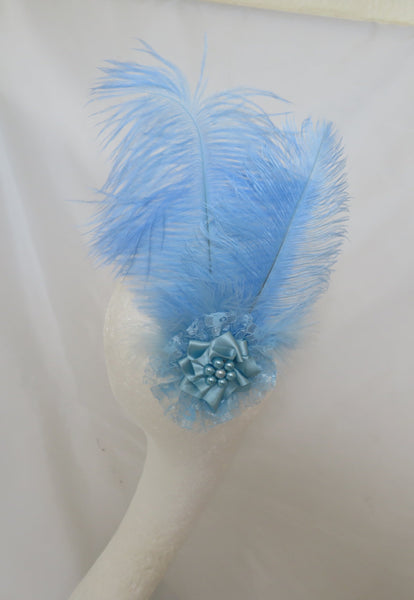 A light blue feather fascinator headpiece with a matching blue fabric flower accent, displayed on a white mannequin head against a white draped backdrop.