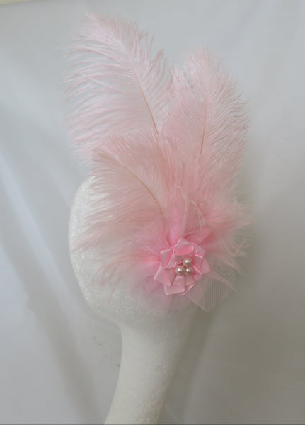 A light pink feather fascinator or hair accessory with a matching pink fabric flower and small pearl-like center, displayed on a white mannequin head against a white draped backdrop.