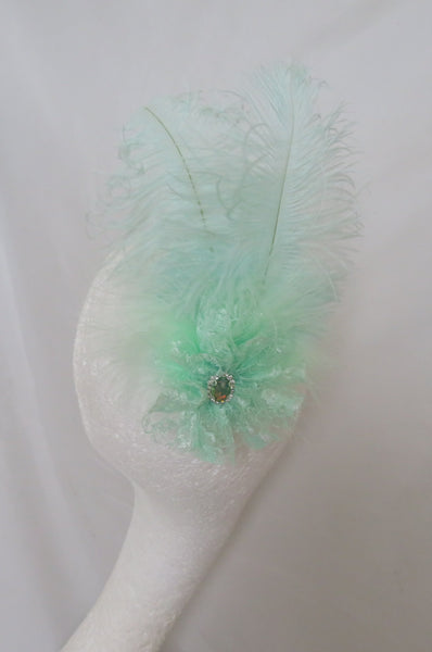 A mint green feather fascinator accessory with a small rhinestone center, displayed on a white mannequin head against a draped white fabric background.