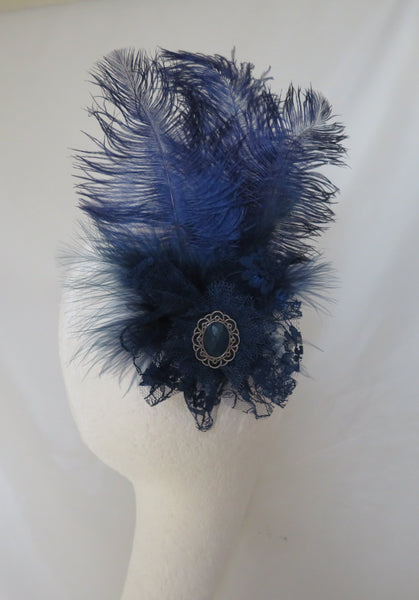 A dark blue feather fascinator headpiece with a lace center and decorative brooch, displayed on a white mannequin head against a white fabric backdrop.