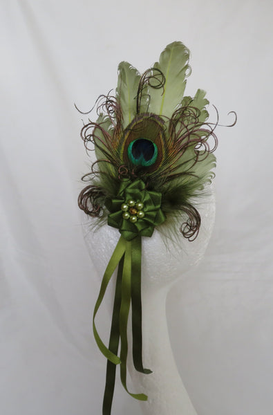 A decorative peacock feather hair accessory displayed on a white mannequin head against a white fabric backdrop. The accessory features multiple green feathers, a prominent peacock feather with a blue-green eye, brown curled feather accents, a green fabric flower, and long olive green ribbons hanging down.
