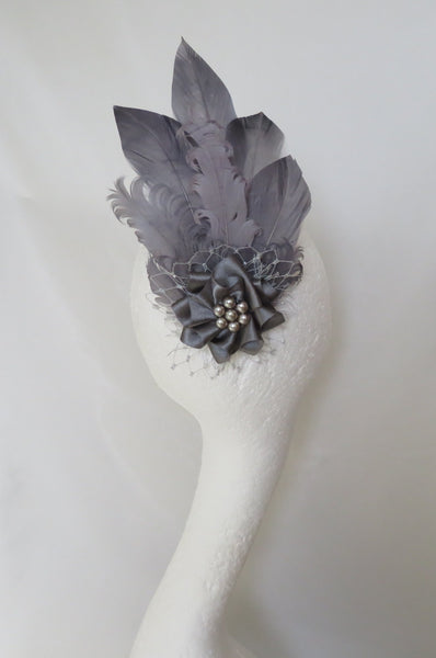 A gray feather fascinator headpiece displayed on a white mannequin head against a draped white fabric background.