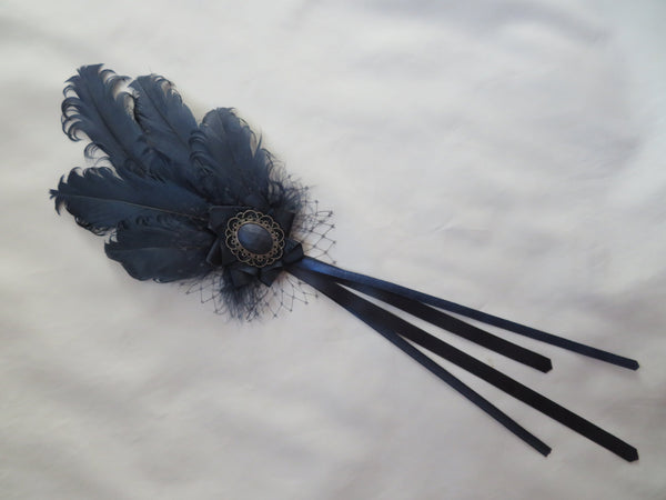 A dark navy blue feather hair accessory with a central decorative brooch and long ribbon tails, laid flat on a white fabric background.