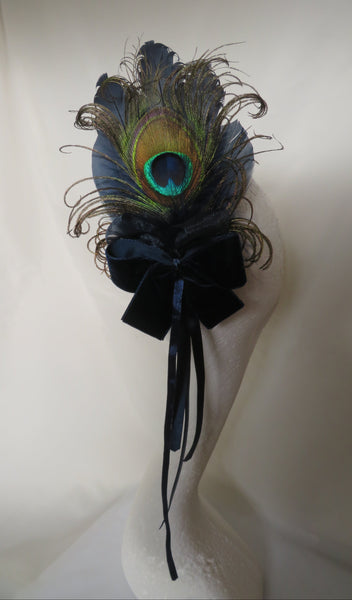 A decorative black velvet bow and ribbon accessory embellished with peacock feathers, displayed on a white mannequin head against a draped white fabric background.
