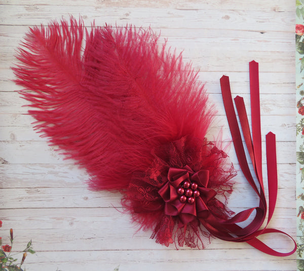 A red feather headband with a large feather, tulle flower, red beads, and ribbon ties, displayed on a light wood background.