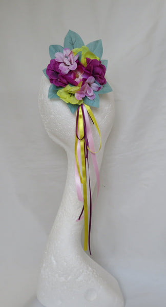 A handmade fabric flower headband with purple, pink, and yellow flowers, teal leaves, and hanging pink, yellow, ribbons, displayed on a white mannequin head against a white fabric backdrop.