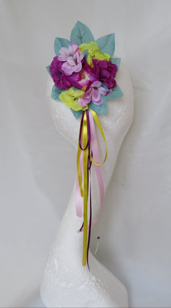 A handmade fabric flower bouquet with purple, pink, and yellow flowers, light blue leaves, and hanging yellow, pink, and brown ribbons, displayed on a white swan head mannequin against a white shower curtain backdrop.