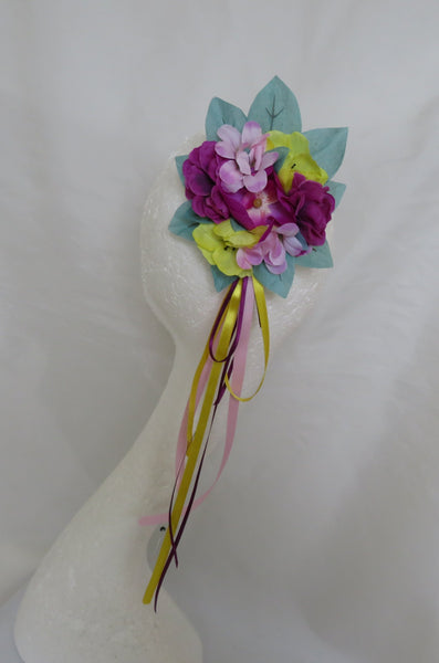 A fabric flower bouquet accessory displayed on a white mannequin torso, featuring purple and pink flowers, green and yellow leaves, and hanging ribbon tails, set against a white draped backdrop 