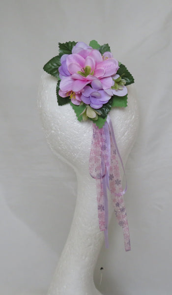 A decorative floral hair accessory with pink and purple artificial flowers, green leaves, and a dangling sheer purple ribbon, displayed on a white mannequin head against a white fabric backdrop.