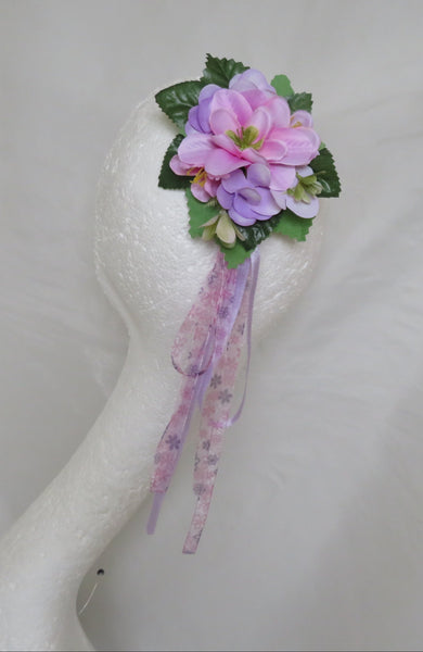 A small floral wristlet or corsage accessory with pink and purple flowers, green leaves, and a sheer purple ribbon, displayed on a white mannequin head against a white fabric backdrop.