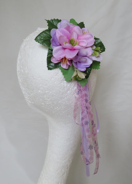 A floral hair accessory featuring pink and purple artificial flowers with green leaves and a dangling sheer purple ribbon, displayed on a white mannequin head against a white fabric backdrop.