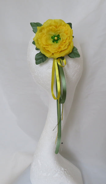A yellow artificial flower with green leaves and a yellow and green ribbon, displayed on a white mannequin head against a white fabric backdrop.