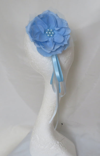 A light blue fabric flower accessory with a small beaded center and a matching blue ribbon, displayed on a white mannequin head against a draped white fabric backdrop.