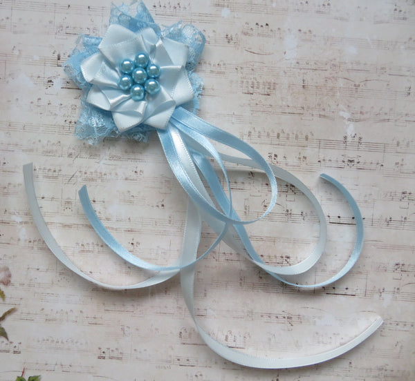 A light blue fabric flower hair accessory with layered tulle, a central cluster of blue beads, and long flowing ribbon tails, displayed on a vintage-style sheet music background.