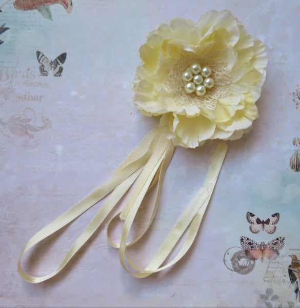 A pale yellow fabric flower hair accessory with a pearl-like center and long matching ribbon tails, laid on a vintage-style floral background.