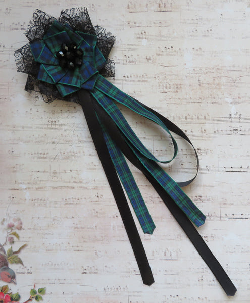 A decorative hair bow accessory featuring a green and black plaid fabric flower with black lace trim and long black ribbon tails, displayed on a vintage-style sheet music background.