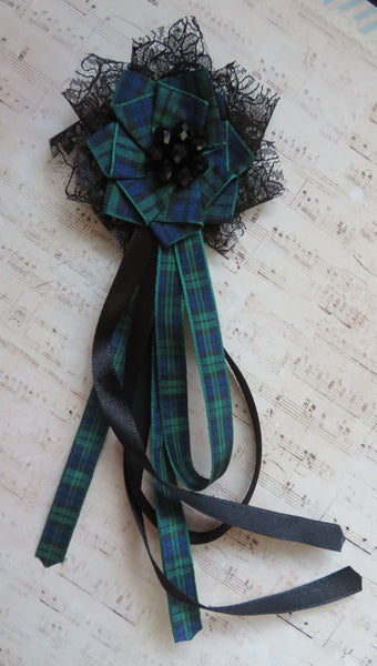 A handmade Scottish tartan brooch featuring a green and blue plaid ribbon flower with black lace, a black beaded center, and long trailing black and plaid ribbons, displayed on a sheet music background.