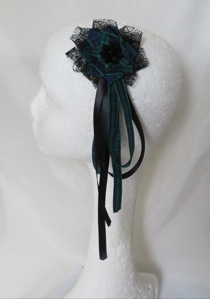 A black lace and tartan floral hair accessory with dark green and black ribbon tails, displayed on a white mannequin head against a white fabric backdrop.