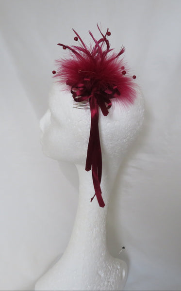 A dark red fascinator hair accessory with feathers and ribbon, displayed on a white mannequin head against a white fabric backdrop.