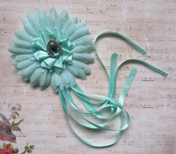 A mint green fabric flower headband with a central rhinestone brooch and long trailing ribbons, displayed on a vintage-style sheet music background.