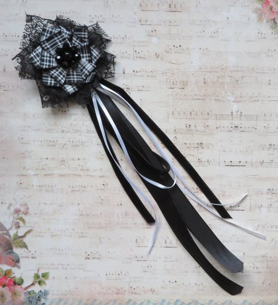 A decorative black and white ribbon brooch with a plaid patterned center and long trailing ribbons, placed on a vintage-style sheet music background with floral accents.